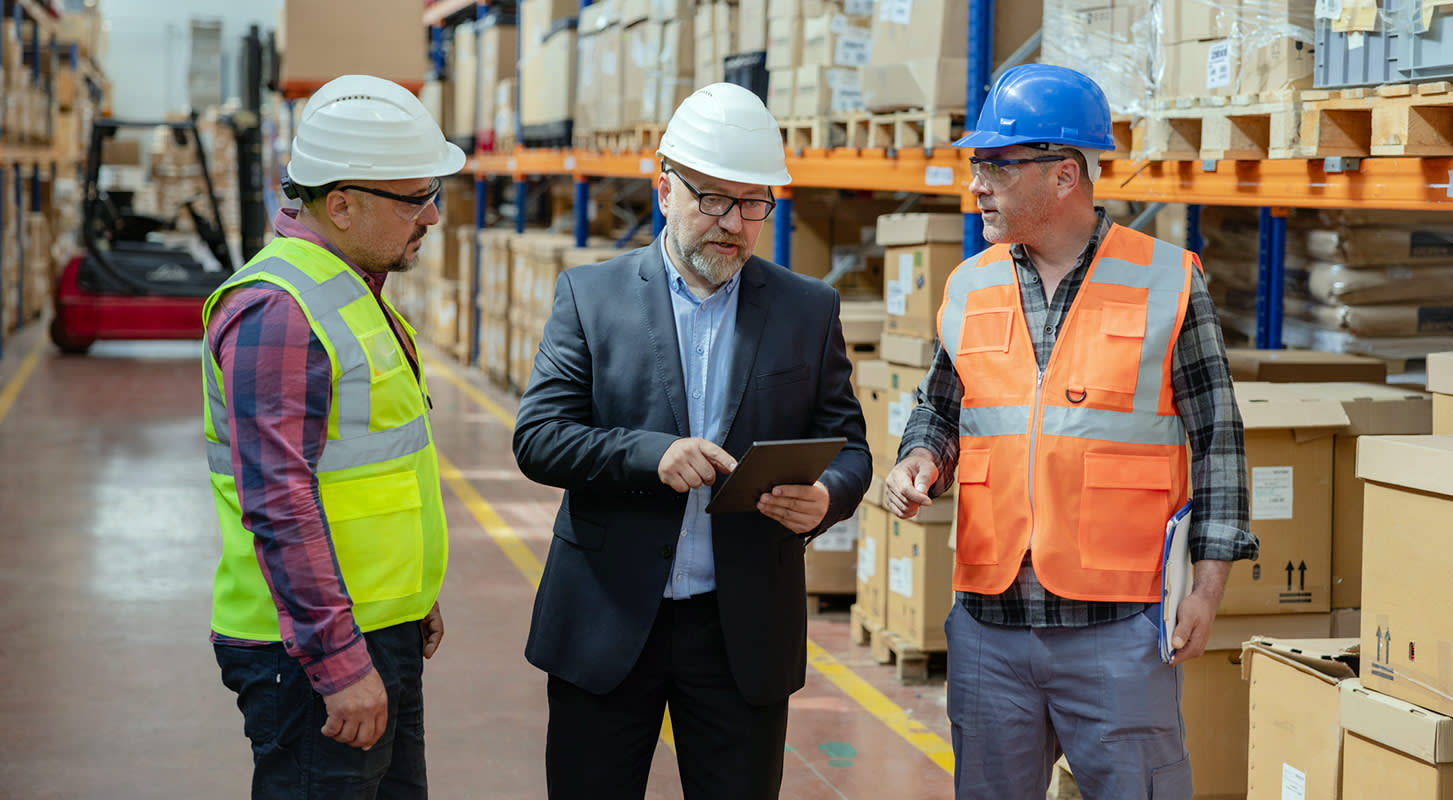 Three men, two in safety vests and hard hats and one in a suit and hard hat, discussing something while looking at a tablet in a warehouse.